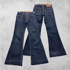Könnte beinhalten: Zwei Jeanshosen mit Schlaghose aus dunkelblauem Denim. Eine Hose hat einen Knopfverschluss, die andere einen Reißverschluss. Beide Hosen haben Front- und Gesäßtaschen und hängen an Holzbügeln.