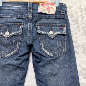 May include: Dark wash denim jeans with a classic five-pocket design. Features include decorative stitching, silver rivets, and a red and white True Religion brand patch. The back pockets have a signature horseshoe design.