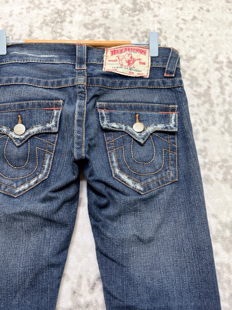 May include: Dark wash denim jeans with a classic five-pocket design. Features include decorative stitching, silver rivets, and a red and white True Religion brand patch. The back pockets have a signature horseshoe design.