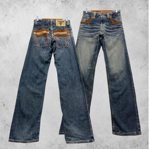 May include: Two pairs of blue denim jeans hanging on wooden hangers. The jeans feature brown suede-like accents on the back pockets and front pockets. A leather patch with a bull logo is visible on the back.