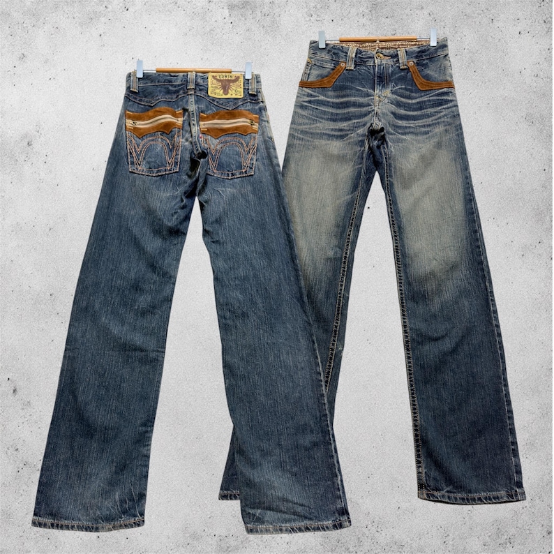 May include: Two pairs of blue denim jeans hanging on wooden hangers. The jeans feature brown suede-like accents on the back pockets and front pockets. A leather patch with a bull logo is visible on the back.