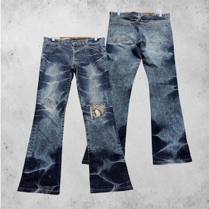 May include: Blue denim jeans with a unique, distressed wash pattern. The jeans feature a flared leg and a patch with a snakeskin design on one knee. The jeans are displayed on a hanger, showcasing both the front and back.