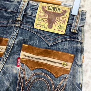 May include: Close-up of vintage Edwin jeans. The blue denim jeans feature brown suede accents on the pockets and a zipper detail. The back pocket has a unique design with decorative stitching. A yellow and brown Edwin label is visible.