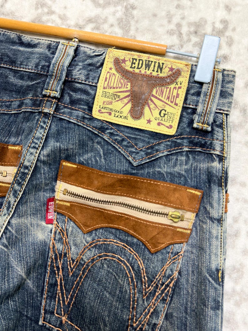 May include: Close-up of vintage Edwin jeans. The blue denim jeans feature brown suede accents on the pockets and a zipper detail. The back pocket has a unique design with decorative stitching. A yellow and brown Edwin label is visible.