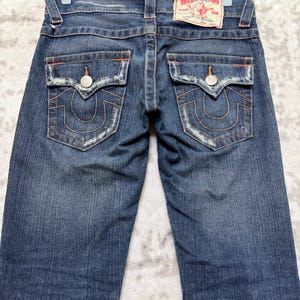 May include: Dark wash denim jeans with decorative stitching and a branded patch. The jeans feature two back pockets with a unique design, metal button closures, and a classic five-pocket style. The jeans are hung on a hanger.