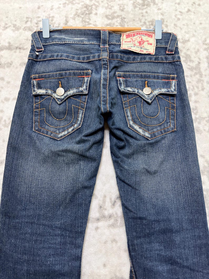 May include: Dark wash denim jeans with decorative stitching and a branded patch. The jeans feature two back pockets with a unique design, metal button closures, and a classic five-pocket style. The jeans are hung on a hanger.