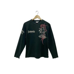 Könnte beinhalten: Dunkelgrünes Langarmshirt mit grafischem Text und Design. Das Shirt zeigt den Text "Tedman's" und ein Design auf der Vorderseite und dem linken Ärmel. Das Design beinhaltet eine rot-weiße Illustration und zusätzlichen Text. Das Shirt hängt an einem Holzbügel.