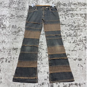 Könnte beinhalten: Blaue und braune Jeans mit einem geschichteten Design. Die Jeans haben ein ausgestelltes Bein und hängen an einem Holzbügel. Der Denim hat einen Used-Look mit ausgefransten Kanten.