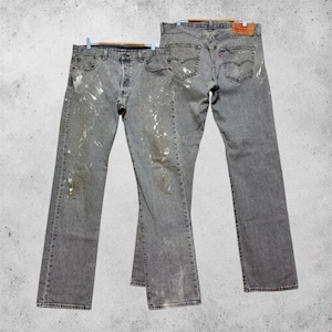Puede incluir: Dos pares de pantalones vaqueros grises con un acabado desgastado y salpicaduras de pintura blanca. Los vaqueros incluyen bragueta de botones, cinco bolsillos y una etiqueta de cuero marrón en la cintura con el logo y texto de Levi's visible.