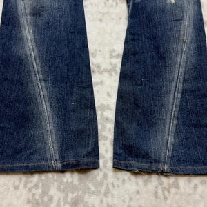 May include: Close-up of the lower portion of a pair of blue denim jeans. The jeans have a flared leg design with a lighter wash detail down the front of each leg. The hem is finished with a brown stitch.