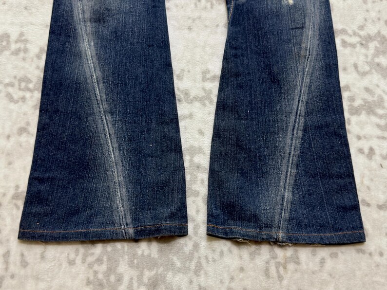 May include: Close-up of the lower portion of a pair of blue denim jeans. The jeans have a flared leg design with a lighter wash detail down the front of each leg. The hem is finished with a brown stitch.
