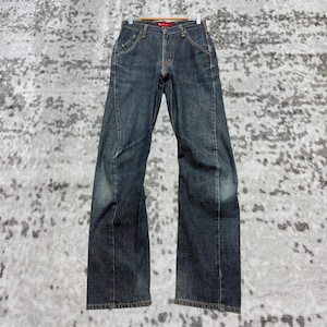 Può includere: Jeans in denim blu scuro con una leggera svasatura. I jeans hanno un classico design a cinque tasche, una chiusura con bottone e cerniera e un'etichetta rossa sul retro. I jeans sono appesi a una gruccia di legno.