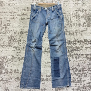 May include: Light blue denim jeans with a distinctive patchwork design. The jeans have multiple panels and a darker blue patch on the lower leg. They are hanging on a wooden hanger, showing their full length and style.