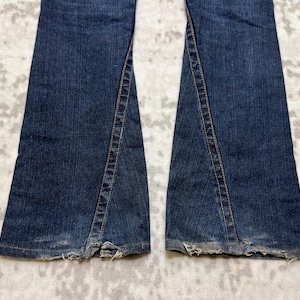 May include: Close-up of the bottom of a pair of dark blue denim jeans. The jeans have a split seam detail on the outer side of each leg, with frayed edges at the hem. The denim has a worn appearance.