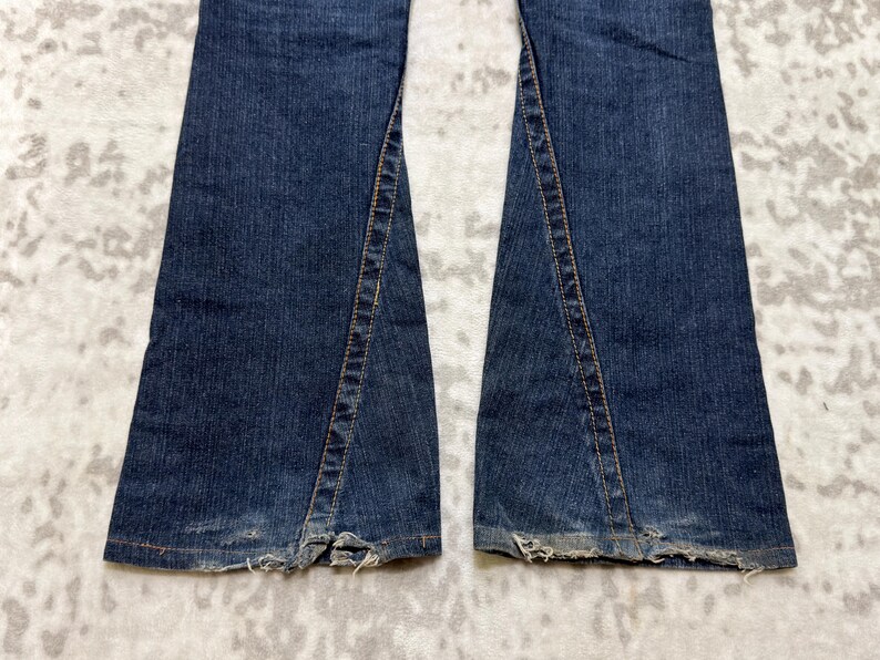 May include: Close-up of the bottom of a pair of dark blue denim jeans. The jeans have a split seam detail on the outer side of each leg, with frayed edges at the hem. The denim has a worn appearance.