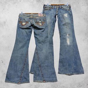 Peut inclure: Deux paires de jeans en denim bleu clair avec une coupe évasée. Les jeans présentent des détails usés, des poches arrière à rabat boutonné avec des surpiqûres orange, et une coupe classique à cinq poches avec fermeture à bouton et braguette.