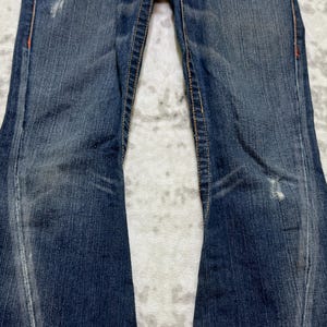 May include: Close-up of blue denim jeans with a slight flare. The jeans show signs of wear with faded areas and distressed details. Orange stitching accents the seams. The fabric has a textured appearance.