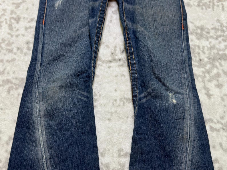 May include: Close-up of blue denim jeans with a slight flare. The jeans show signs of wear with faded areas and distressed details. Orange stitching accents the seams. The fabric has a textured appearance.