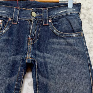 May include: Close-up of dark wash denim jeans with distressed details. The jeans feature a button closure, front pockets, and decorative stitching in orange and red. The jeans are hanging on a wooden hanger.