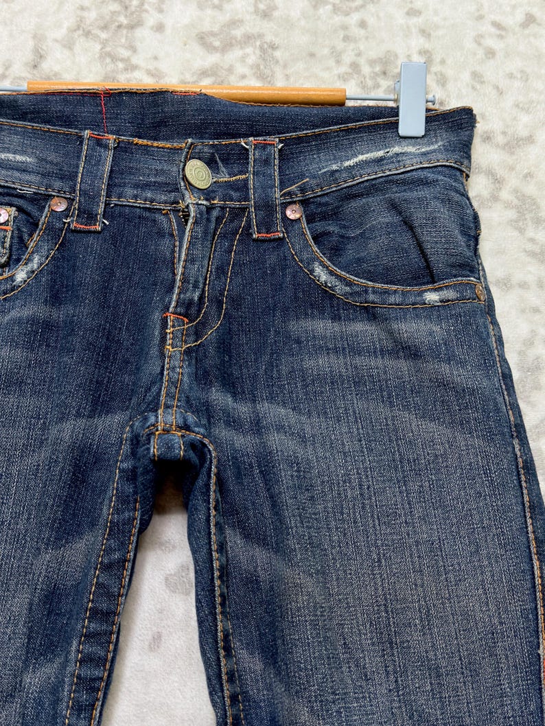 May include: Close-up of dark wash denim jeans with distressed details. The jeans feature a button closure, front pockets, and decorative stitching in orange and red. The jeans are hanging on a wooden hanger.