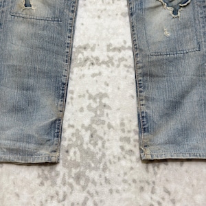Puede incluir: Vaqueros de mezclilla azul descolorido con detalles desgastados, incluyendo rasgaduras y parches. Los vaqueros tienen un dise&ntilde;o cl&aacute;sico de cinco bolsillos y un corte de pierna recta. La mezclilla tiene un aspecto vintage.