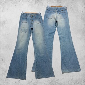 Puede incluir: Un par de pantalones vaqueros de campana en azul lavado claro con diseño de cintura alta, cierre de botones, dos bolsillos delanteros y un pequeño detalle de rosa bordada en la parte superior del muslo, vistos de frente y de espaldas.