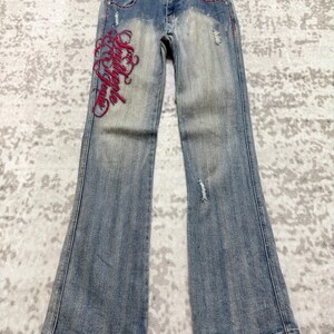 May include: Light blue denim jeans with a flared leg. The jeans feature red stitching and distressed details, including a small rip on the right leg. The left leg has red embroidered script. The jeans are hanging on a wooden hanger.