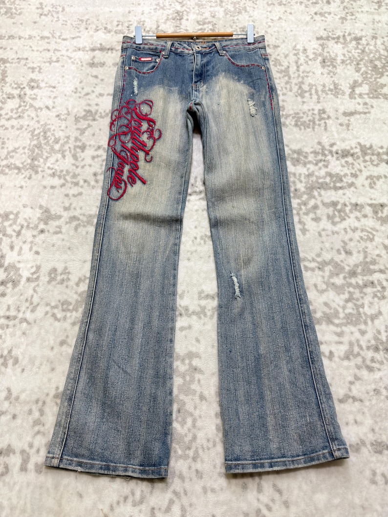 May include: Light blue denim jeans with a flared leg. The jeans feature red stitching and distressed details, including a small rip on the right leg. The left leg has red embroidered script. The jeans are hanging on a wooden hanger.