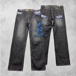 Könnte beinhalten: Eine dunkelgraue Jeans aus Denim mit einem blauen, gestickten Drachenmotiv auf dem linken Vorderbein, Akzenten aus blau-weiß gemustertem Stoff an den Vorder- und Gesäßtaschen, einem Knopfverschluss und Gürtelschlaufen am Bund.