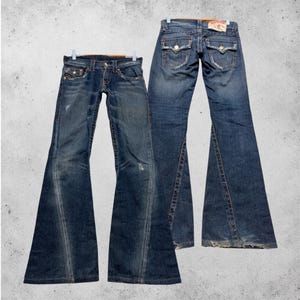 May include: Two pairs of flared-leg blue denim jeans. The jeans are displayed on hangers against a gray background. The jeans have a distressed look with visible wear and tear, and feature classic five-pocket styling.
