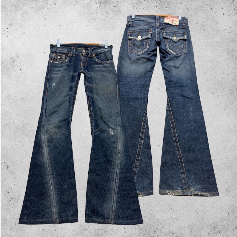 May include: Two pairs of flared-leg blue denim jeans. The jeans are displayed on hangers against a gray background. The jeans have a distressed look with visible wear and tear, and feature classic five-pocket styling.