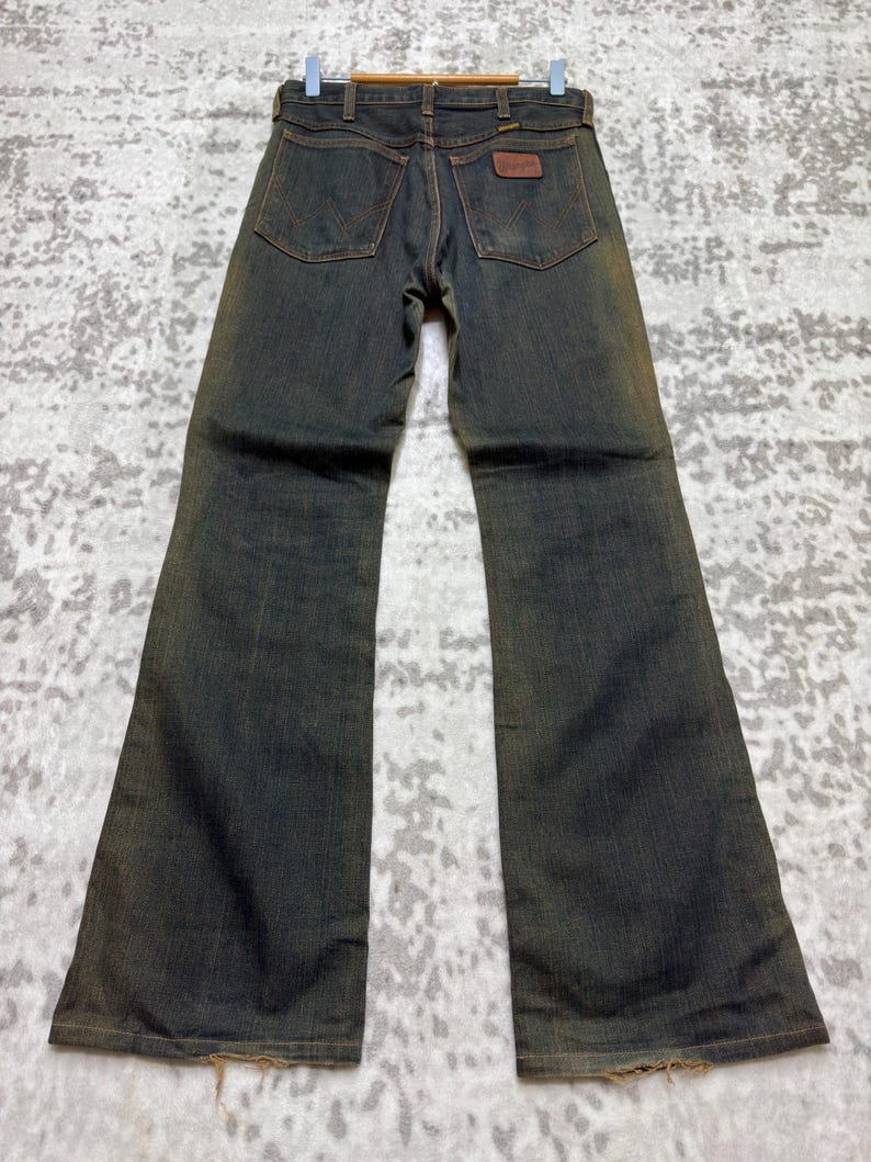 May include: Vintage denim jeans with a flared leg design. The jeans are a dark wash with a faded, worn appearance. Features include classic five-pocket styling, a brown leather patch, and a metal zipper. The jeans are displayed on a hanger.