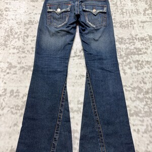 May include: Dark wash denim jeans with a flared leg design. The jeans feature distressed details, orange stitching, and two back pockets with decorative stitching. A leather patch is visible at the waistband.