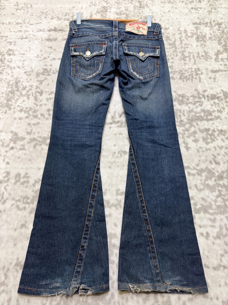 May include: Dark wash denim jeans with a flared leg design. The jeans feature distressed details, orange stitching, and two back pockets with decorative stitching. A leather patch is visible at the waistband.