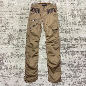 May include: Khaki-colored pants with multiple zippers and decorative stitching. The pants feature a cargo pocket and gathered fabric details. The waistband has colorful accents and the text "ROCK STAR".