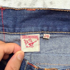 May include: Close-up of blue denim jeans with orange stitching. A white label with the text "TRUE RELIGION" and a graphic of a Buddha playing a guitar is visible. The size tag reads "26".