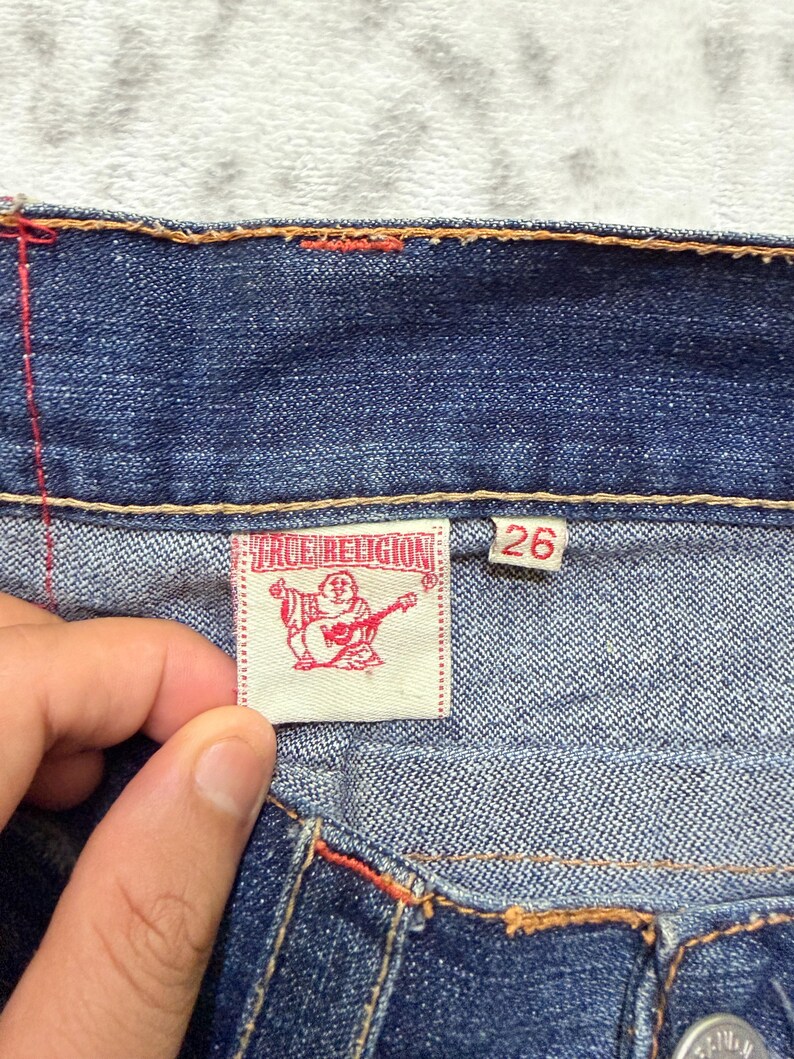 May include: Close-up of blue denim jeans with orange stitching. A white label with the text "TRUE RELIGION" and a graphic of a Buddha playing a guitar is visible. The size tag reads "26".