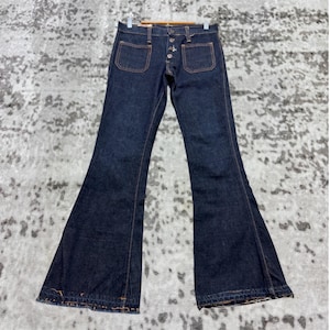 Könnte beinhalten: Dunkelblaue Schlaghose aus Denim mit orangefarbenen Nähten. Die Jeans hat zwei Vordertaschen und einen Knopfverschluss. Die Jeans hängt an einem Holzbügel.
