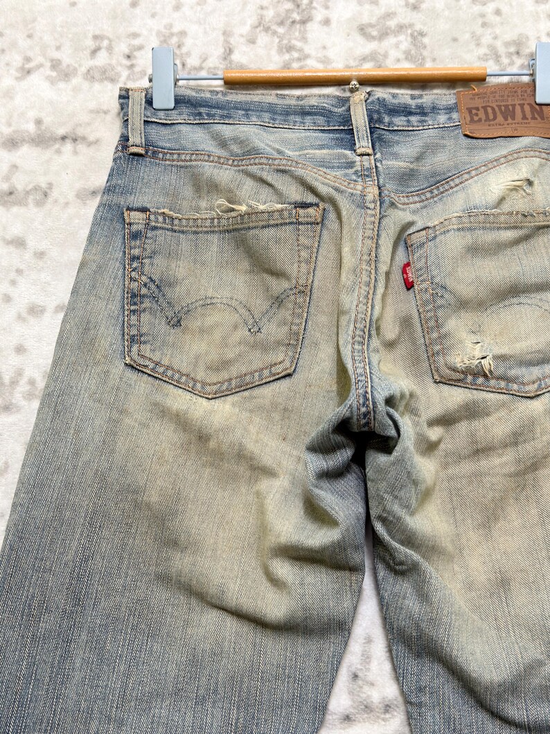 Puede incluir: Vaqueros de mezclilla azul descolorido con detalles desgastados, incluyendo rasgaduras y parches. Los bolsillos traseros presentan un dise&ntilde;o cl&aacute;sico, y una peque&ntilde;a etiqueta roja es visible. Los vaqueros est&aacute;n colgados en una percha de madera, mostrando el estilo vintage.