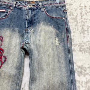 May include: Faded blue denim jeans with red stitching and decorative embroidery. The jeans have a distressed look with a few small tears and a light wash effect. The word "eagle" is visible on the waistband.