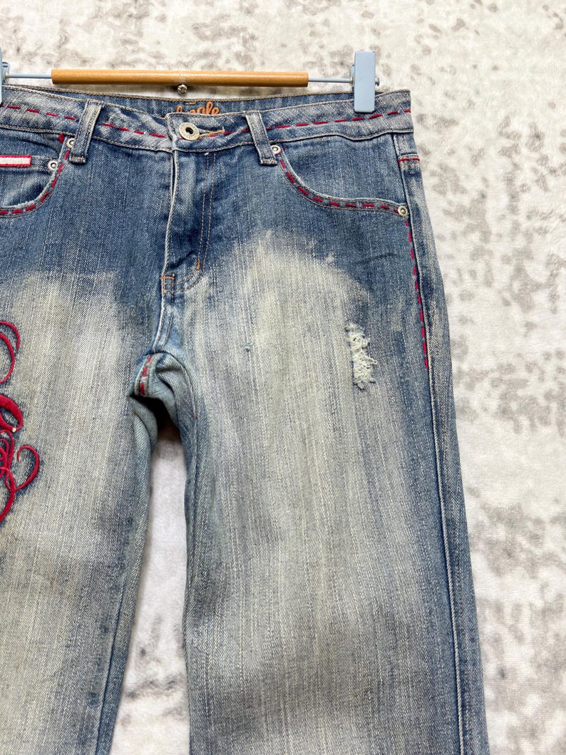 May include: Faded blue denim jeans with red stitching and decorative embroidery. The jeans have a distressed look with a few small tears and a light wash effect. The word "eagle" is visible on the waistband.