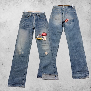 May include: Light blue denim jeans with patches, displayed front and back. The jeans have a distressed look with a hole on the front. Patches include text and graphics. The jeans are hanging on a wooden hanger.
