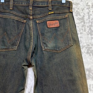 May include: Dark wash denim jeans with brown stitching and a brown leather Wrangler patch on the back pocket. The jeans are hanging on a wooden hanger. The Wrangler logo is visible on the back pocket and the waistband.