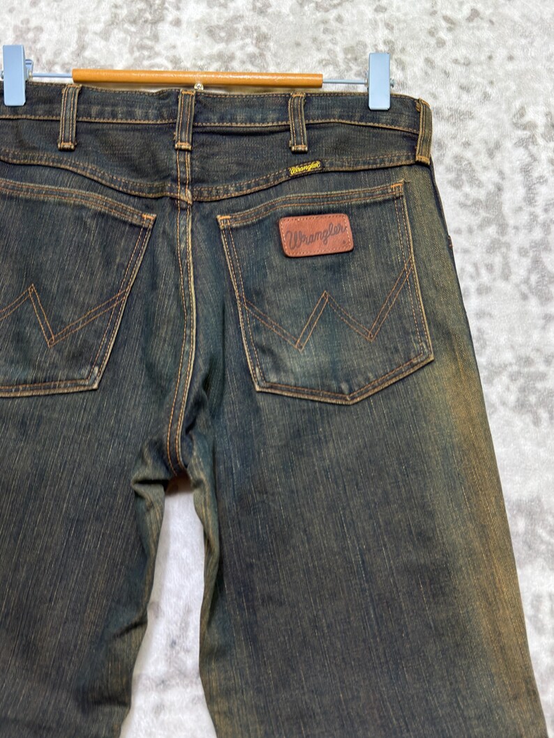 May include: Dark wash denim jeans with brown stitching and a brown leather Wrangler patch on the back pocket. The jeans are hanging on a wooden hanger. The Wrangler logo is visible on the back pocket and the waistband.