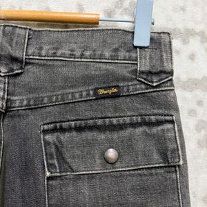 May include: Close-up of a pair of black denim Wrangler jeans. The jeans have a pocket with a silver button and the Wrangler logo is visible. The jeans are hanging on a wooden and metal hanger.