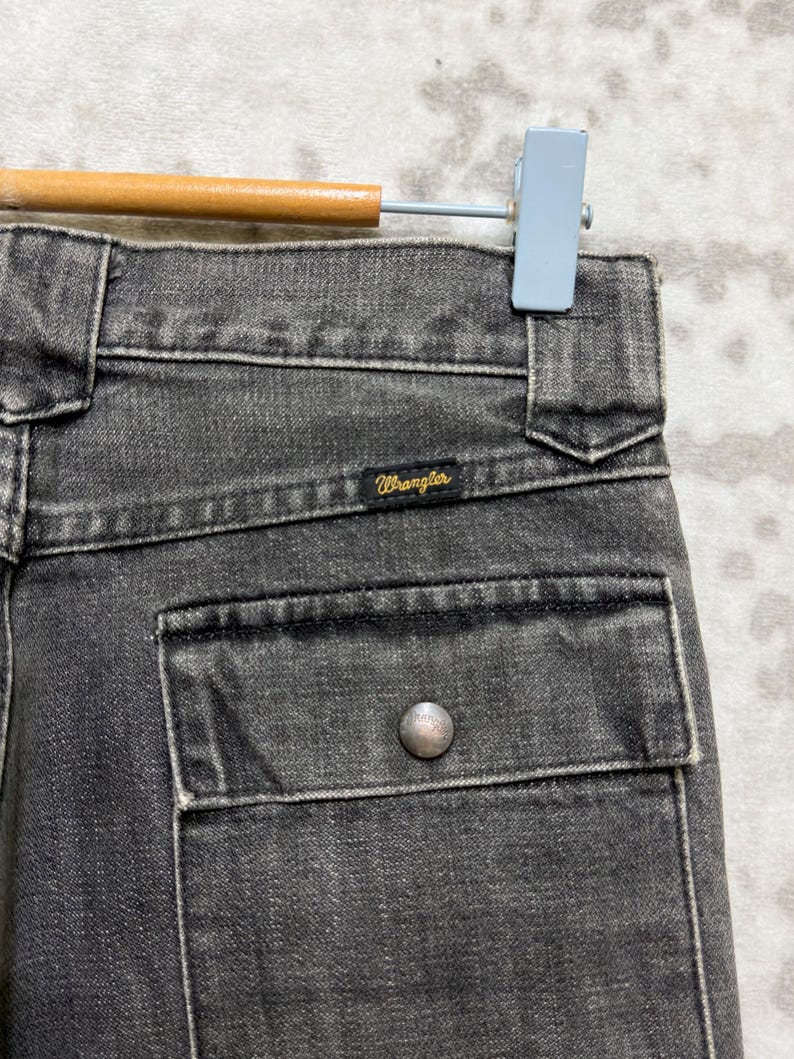 May include: Close-up of a pair of black denim Wrangler jeans. The jeans have a pocket with a silver button and the Wrangler logo is visible. The jeans are hanging on a wooden and metal hanger.