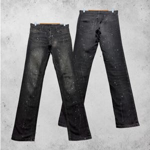 Könnte beinhalten: Zwei Paar schwarze Jeans mit Farbspritzern. Die Jeans werden auf Holzbügeln vor einem neutralen Hintergrund präsentiert. Sie haben ein klassisches Fünf-Taschen-Design und einen Knopfverschluss.