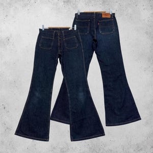 Könnte beinhalten: Zwei Paar dunkelblaue Jeans aus Denim mit ausgestelltem Bein. Das vordere Paar hat zwei große aufgesetzte Taschen und einen Knopfverschluss. Das hintere Paar zeigt zwei Gesäßtaschen, ein kleines rotes Etikett und einen braunen Lederaufnäher.