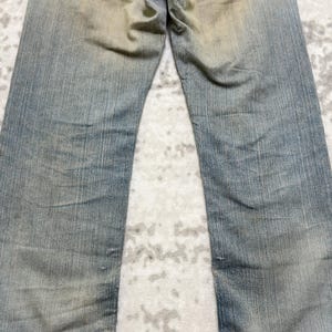 Puede incluir: Vaqueros de mezclilla azul descolorido con corte recto. Los vaqueros muestran signos de desgaste con parches m&aacute;s claros y sutiles desgastes en toda la tela. La apariencia general sugiere una est&eacute;tica vintage o desgastada.
