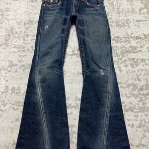 May include: Dark wash denim jeans with a flared leg design. The jeans feature a classic five-pocket style, red stitching, and a button and zipper closure. The jeans are hanging on a white hanger.